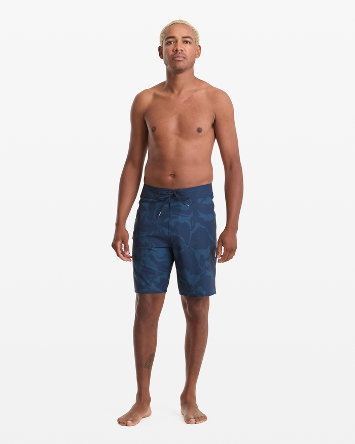 A man with short blond hair stands barefoot against a white background, wearing Volcom Men's Shoreline Mod 20 blue floral swim trunks. He faces forward with a neutral expression and his arms relaxed at his sides.