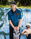 A man in a Volcom Men's Slab City Short Sleeve Tee and black cap leans on a skateboard by a graffiti wall, while another shirtless man in a white cap and sunglasses listens to earphones nearby.