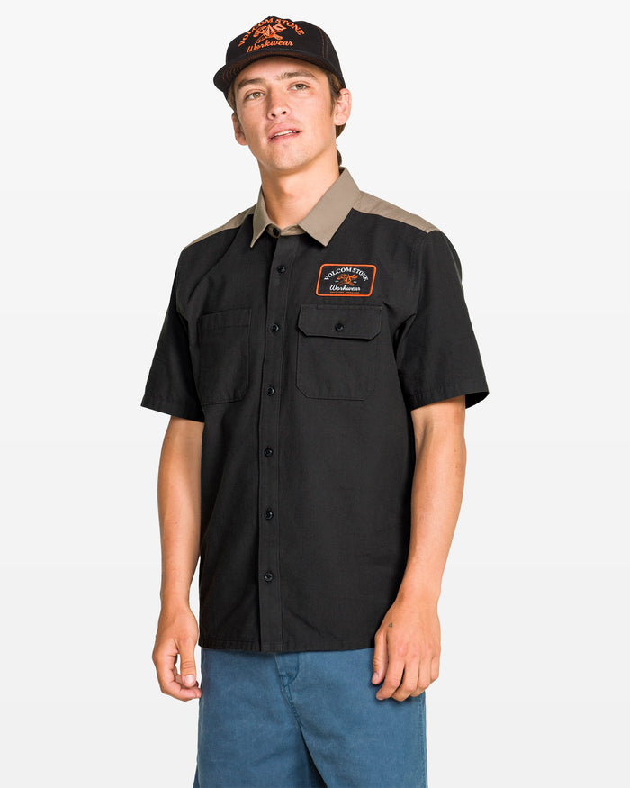 A young man wearing a Volcom Men's Slab City Short Sleeve Tee, blue shorts, and a black cap stands against a plain white background.
