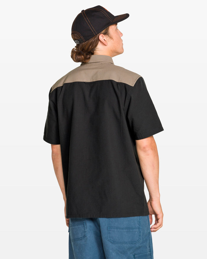 A person with long hair in a ponytail, wearing the Volcom Men's Slab City Short Sleeve Tee, blue pants, and a black cap stands with their back to the camera against a plain background.
