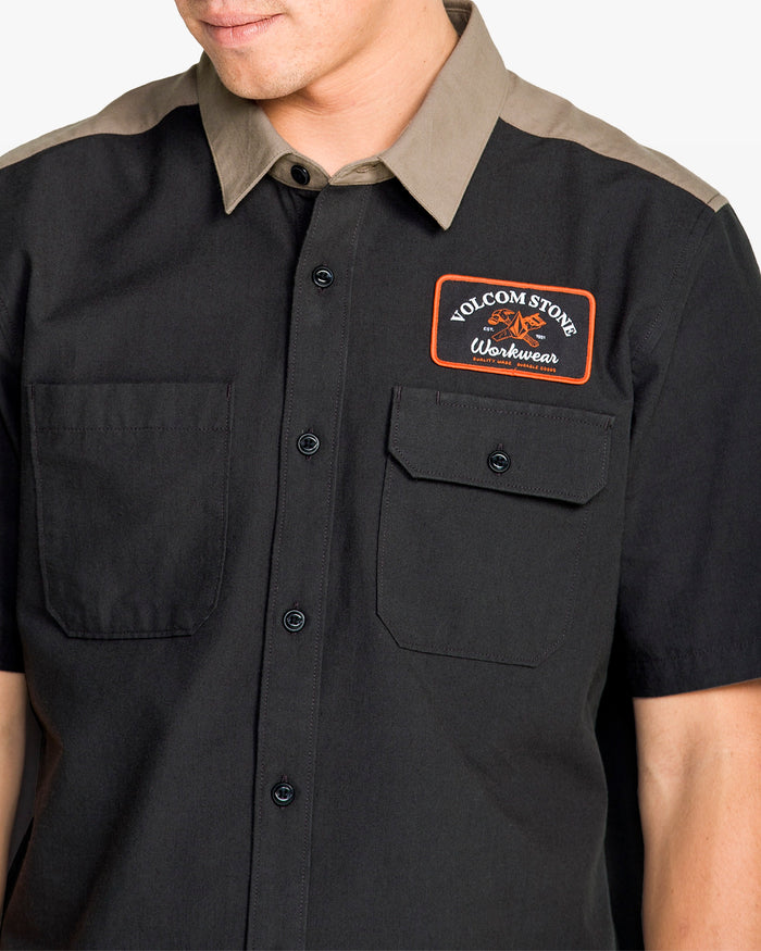 A person wears the Volcom Men's Slab City Short Sleeve Tee, a classic fit two-tone shirt with a tan collar, black body, and a Volcom Stone Workwear patch above the left chest pocket.