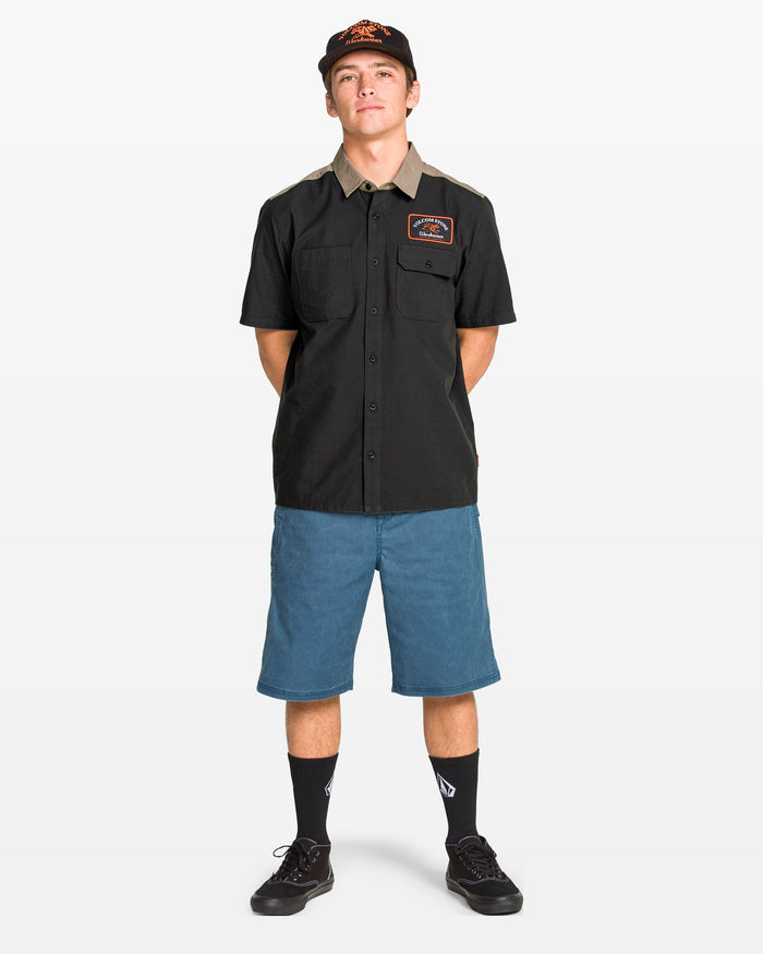 A young man stands facing forward in a classic fit black Volcom Men's Slab City Short Sleeve Tee, blue knee-length shorts, black socks, black shoes, and a black cap, set against a plain white background.