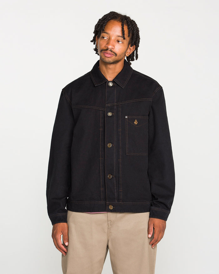 A person with short dreadlocks wears the Volcom Men's Slakers Jacket, a black lightweight piece featuring visible stitching and a small chest pocket, paired with beige pants while standing against a plain white background.
