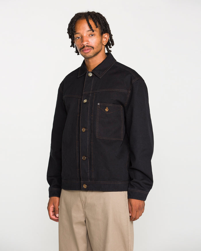 A man with short dreadlocks stands facing forward against a plain white background. He is wearing the Volcom Men's Slakers Jacket—a black cotton canvas jacket featuring brown stitching and a chest pocket—paired with beige pants.
