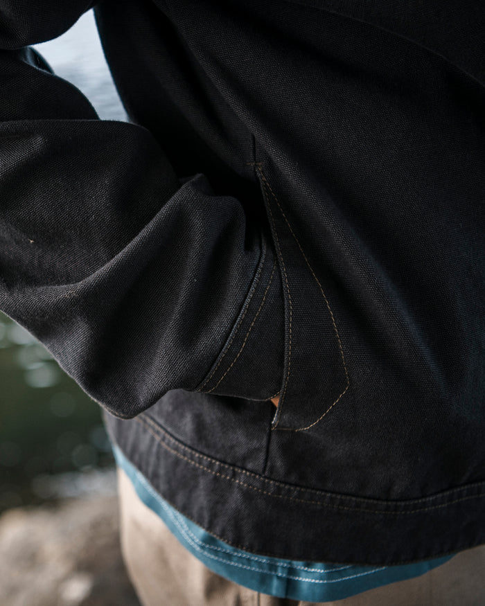 A man wearing the Volcom Men’s Slakers Jacket in a dark cotton canvas stands outdoors near water, his hand tucked into the pocket. Only his torso and arm are visible.
