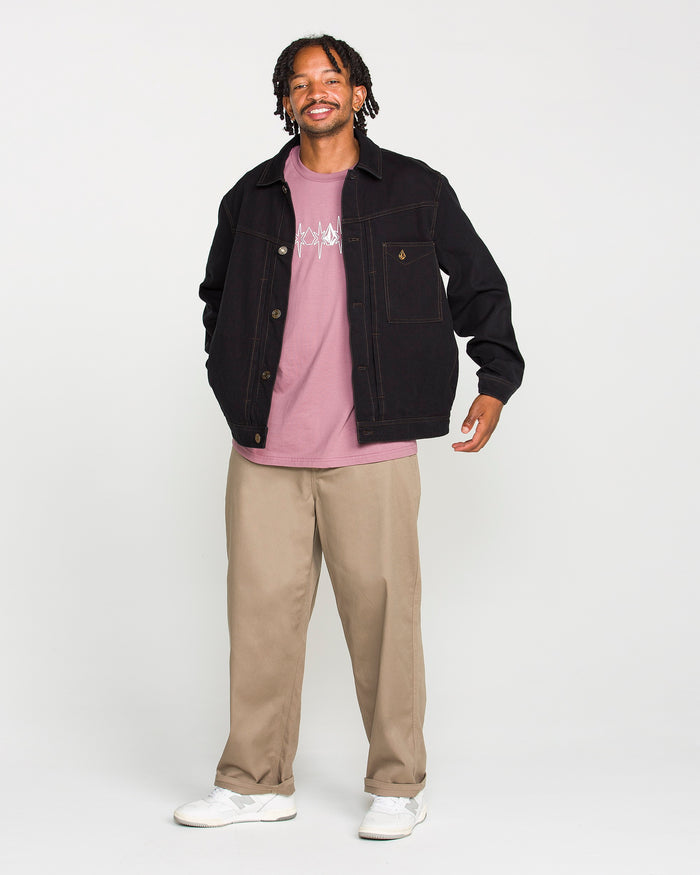A Black man with dreadlocks smiles while modeling the Volcom Men's Slakers Jacket over a pink graphic T-shirt, tan trousers, and white sneakers against a plain light gray background.