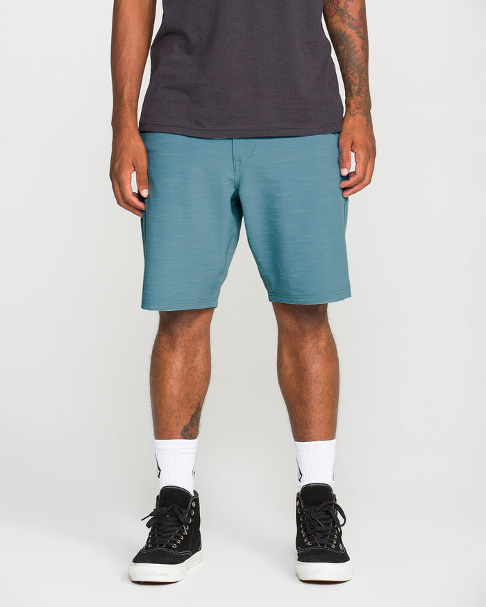 A man is seen from the waist down wearing Volcom Men's Slub Frickin Cross Shred Hybrid Shorts in teal with a modern fit, paired with a black t-shirt, white Nike socks, and black sneakers, standing against a plain light background.