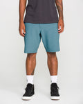 A man is seen from the waist down wearing Volcom Men's Slub Frickin Cross Shred Hybrid Shorts in teal with a modern fit, paired with a black t-shirt, white Nike socks, and black sneakers, standing against a plain light background.