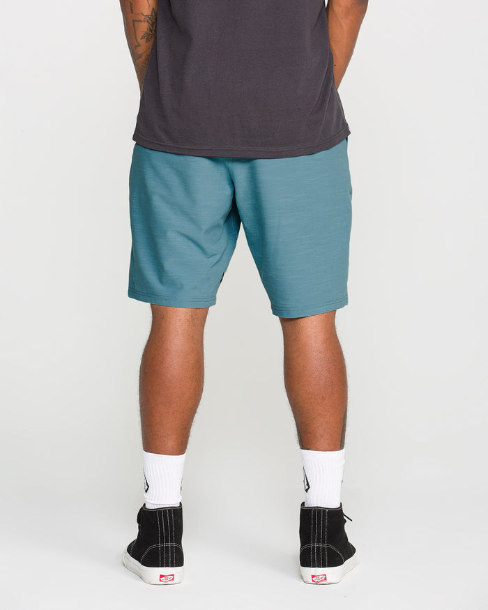 A person stands facing away, wearing a dark t-shirt paired with Volcom Men's Slub Frickin Cross Shred Hybrid Shorts in teal, which feature 4-way stretch for modern fit and comfort. Their outfit is complemented by white crew socks and black sneakers, with only their lower body visible.
