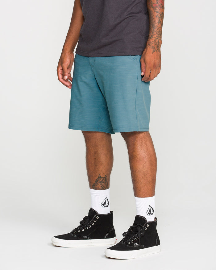 A person wearing Volcom Men's Slub Frickin Cross Shred Hybrid Shorts in teal with a modern fit, a dark T-shirt, white socks featuring a black logo, and black high-top sneakers stands against a plain white background.