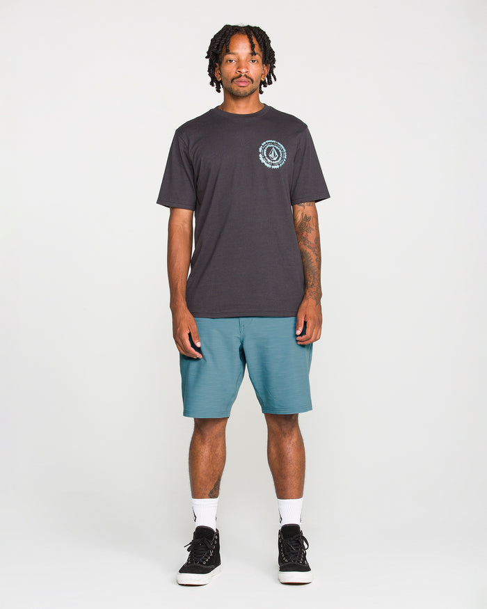 A man with short dreadlocks and visible arm tattoos stands against a plain white background, wearing a black T-shirt with a circular logo on the chest, Volcom Men's Slub Frickin Cross Shred Hybrid Shorts in teal, white socks, and black sneakers.