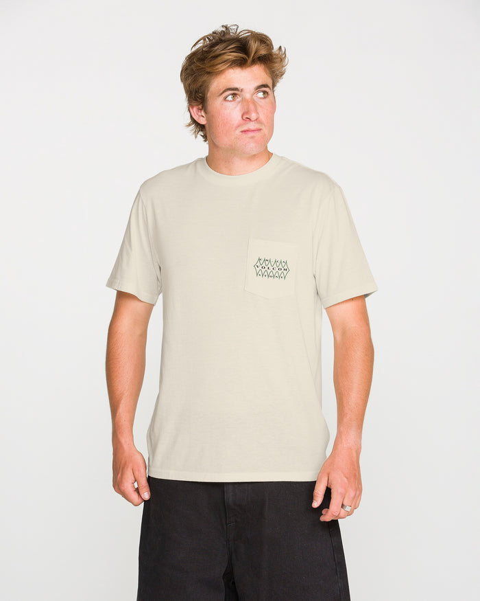 A person is wearing the Volcom Men's Smokers Lounge Short Sleeve Tee—a light beige crew neck t-shirt featuring a small geometric design on the pocket—paired with black shorts, standing against a plain white background and looking slightly to their right.