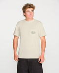 A person is wearing the Volcom Men's Smokers Lounge Short Sleeve Tee—a light beige crew neck t-shirt featuring a small geometric design on the pocket—paired with black shorts, standing against a plain white background and looking slightly to their right.