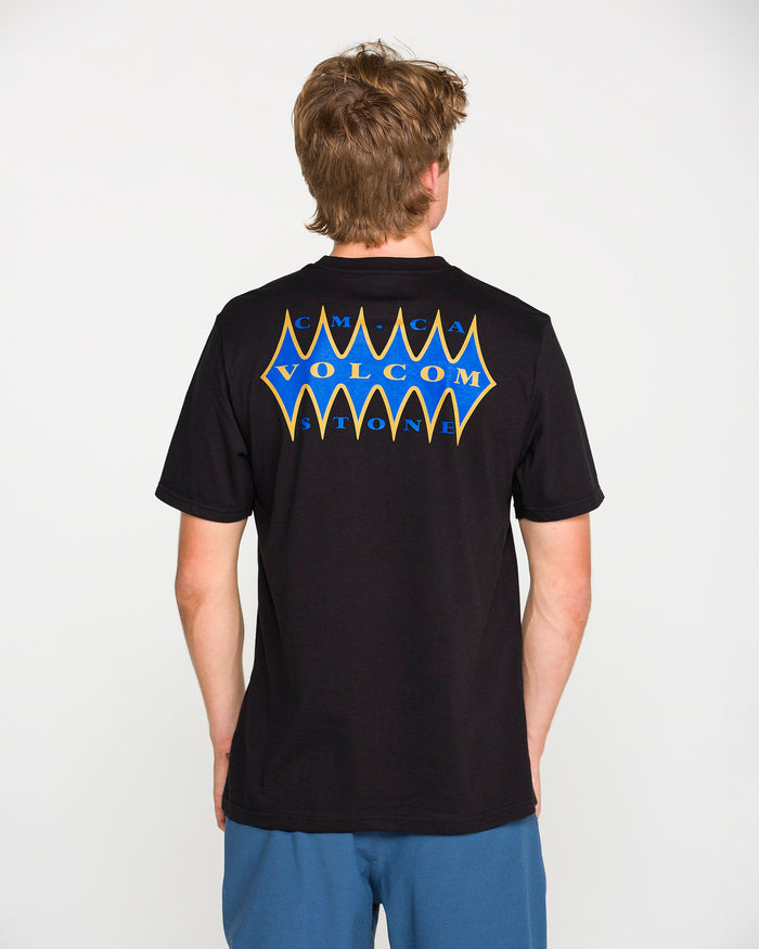 A person with short brown hair, wearing the Volcom Men's Smokers Lounge Short Sleeve Tee in washed black heather—with a blue and yellow Volcom Stone graphic on the back—stands facing away from the camera.