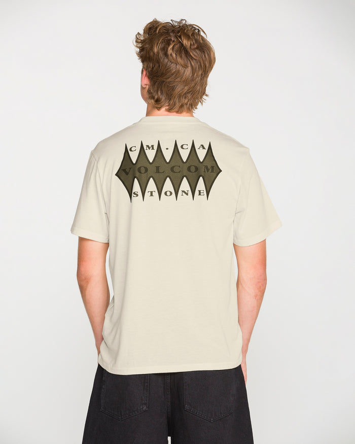 A person wearing the Volcom Men's Smokers Lounge Short Sleeve Tee—a modern fit, light-colored crew neck featuring a large black bat-shaped graphic and the words “CMCA VOLCOM STONE” on the back—stands against a plain white background.