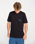 A person with light brown hair looks down while wearing the Volcom Men's Smokers Lounge Short Sleeve Tee, featuring a patterned chest pocket, and blue shorts, standing against a plain white background.