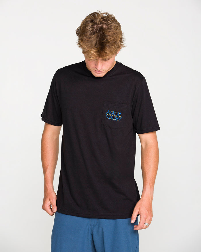 A person with light brown hair looks down while wearing the Volcom Men's Smokers Lounge Short Sleeve Tee, featuring a patterned chest pocket, and blue shorts, standing against a plain white background.