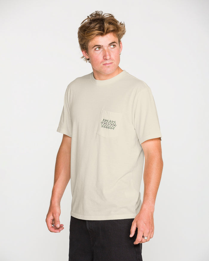 A young man with tousled hair wears the Volcom Men's Smokers Lounge Short Sleeve Tee in light beige, featuring a small black design on the chest pocket, paired with black pants. He stands against a plain white background, looking slightly to his left.