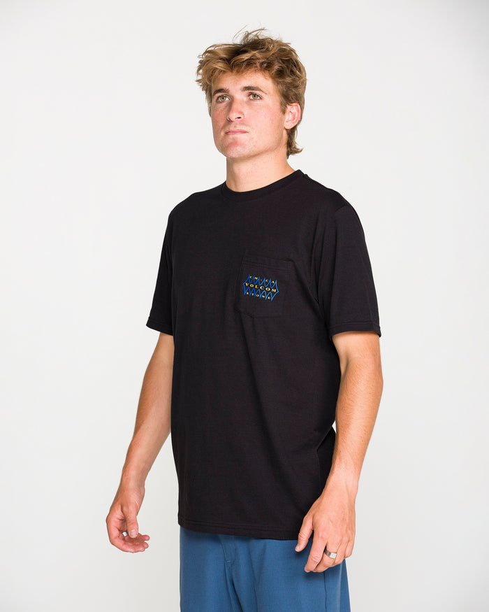 A young man with tousled hair wears the Volcom Men's Smokers Lounge Short Sleeve Tee in washed black heather, featuring a small chest pocket with a subtle pattern. He pairs it with blue pants and stands against a plain white background, looking slightly upward and to the side.