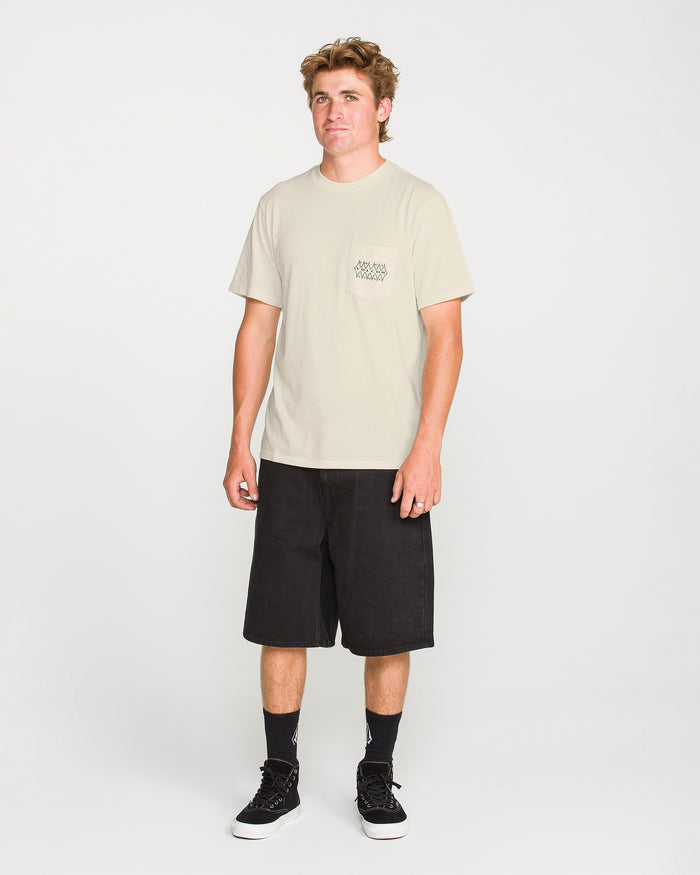 A person stands facing forward wearing the Volcom Men's Smokers Lounge Short Sleeve Tee in light beige, paired with black knee-length shorts, black crew socks, and black sneakers against a plain white background.