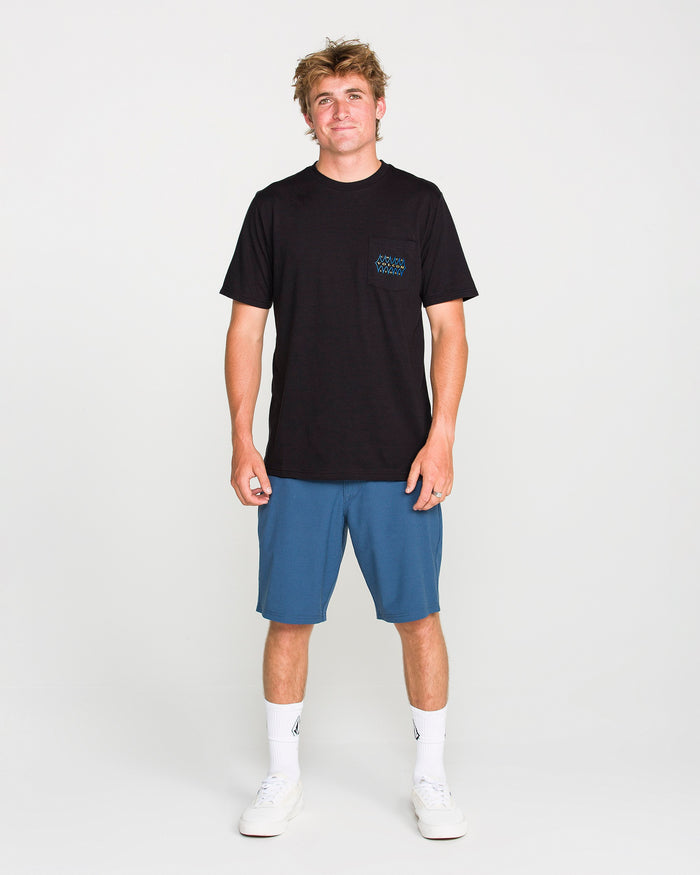 A young man stands facing forward and smiling against a plain white background, wearing the Volcom Men's Smokers Lounge Short Sleeve Tee in washed black heather with a patterned chest pocket, paired with blue shorts, white socks, and white sneakers.