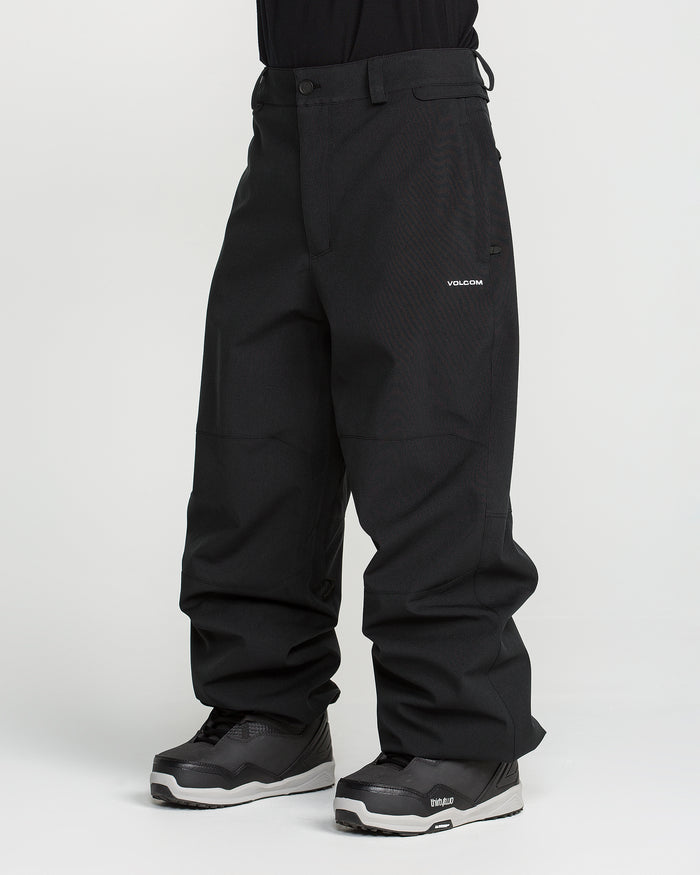 A person wearing loose-fitting black Volcom Men’s Snow Billow Pants, made with 10K waterproof V-Science 2-Layer fabric, and gray-black snow boots stands against a plain white background. Only the lower body and legs are visible; the upper body is cropped out.