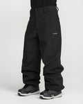 A person wearing loose-fitting black Volcom Men’s Snow Billow Pants, made with 10K waterproof V-Science 2-Layer fabric, and gray-black snow boots stands against a plain white background. Only the lower body and legs are visible; the upper body is cropped out.