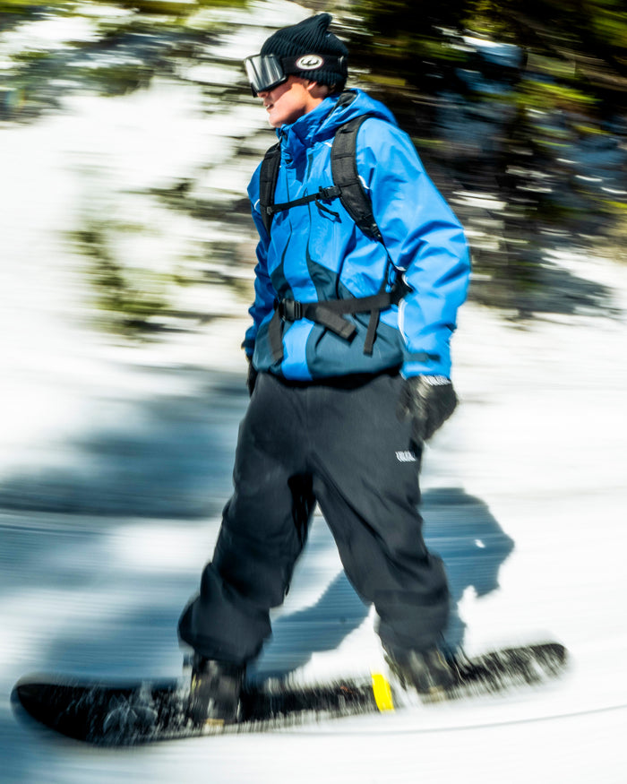 Wearing a blue jacket, black pants, and goggles—outfitted with Volcom Men’s Snow Billow Pants featuring 10K waterproof protection—a snowboarder races swiftly down a snowy slope, trees blurred in the background.