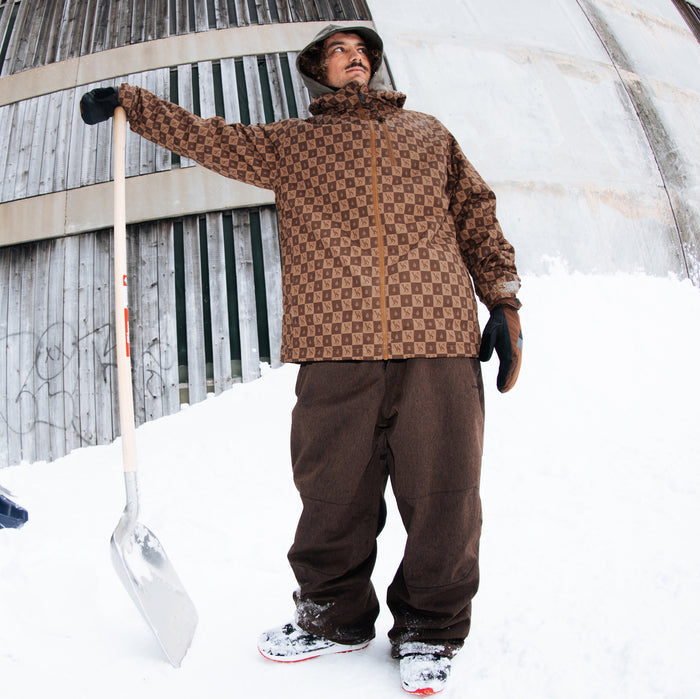 A man wearing a brown and tan checkered winter jacket, brown snow pants, and a hat stands in the snow holding a shovel. The background shows a snowy outdoor setting with a wooden fence and a curved concrete wall.