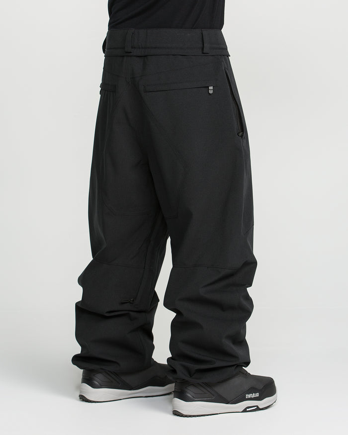A person is shown standing sideways against a plain white background, wearing Volcom Men’s Snow Billow Pants in black and matching black sneakers. The upper body is out of frame. These men’s snow pants feature zippered pockets, reinforced stitching, and are crafted with V-Science 2-Layer fabric for reliable 10K waterproof protection.