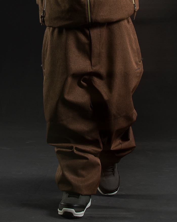 A person wearing loose-fitting brown Volcom Men’s Snow Billow Pants and a matching jacket walks forward, with only their legs and feet visible against a dark background, showcasing the V-Science 2-Layer construction and 10K/10K performance design.