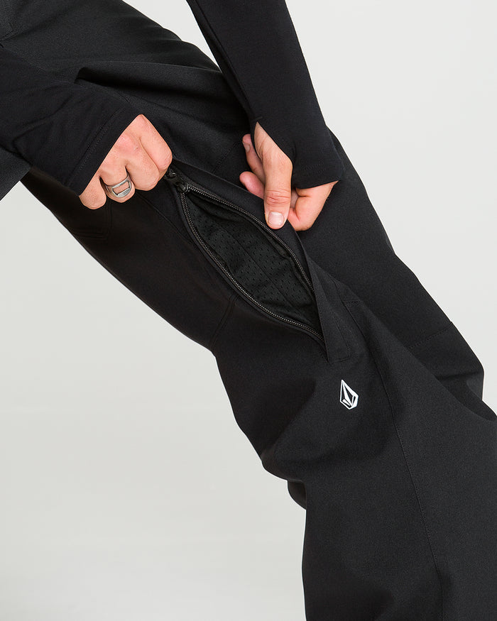 A person unzips a vent on the side of black Volcom Men’s Snow Billow Pants, revealing mesh fabric inside. Crafted with V-Science 2-Layer material and boasting 10K waterproof protection, the pants feature a small, white geometric logo near the knee.