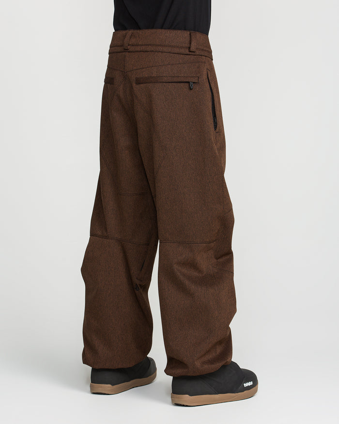 A person is shown from the waist down wearing Volcom Men’s Snow Billow Pants in brown with black boots, standing against a white background. The pants have zipper pockets, seam detailing at the knees, and an adjustable waist.