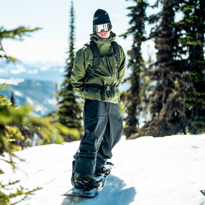 A person in a green jacket, wearing Volcom Men’s Snow Billow Pants featuring V-Science 2-Layer construction and 10K waterproof protection, along with goggles, stands on a snowboard in a snowy mountain landscape surrounded by pine trees.