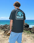 A person with curly hair and sunglasses, wearing a modern fit black Volcom Men's Solstice Short Sleeve Tee and denim shorts, stands on a rocky coastline overlooking the ocean beneath a clear blue sky.