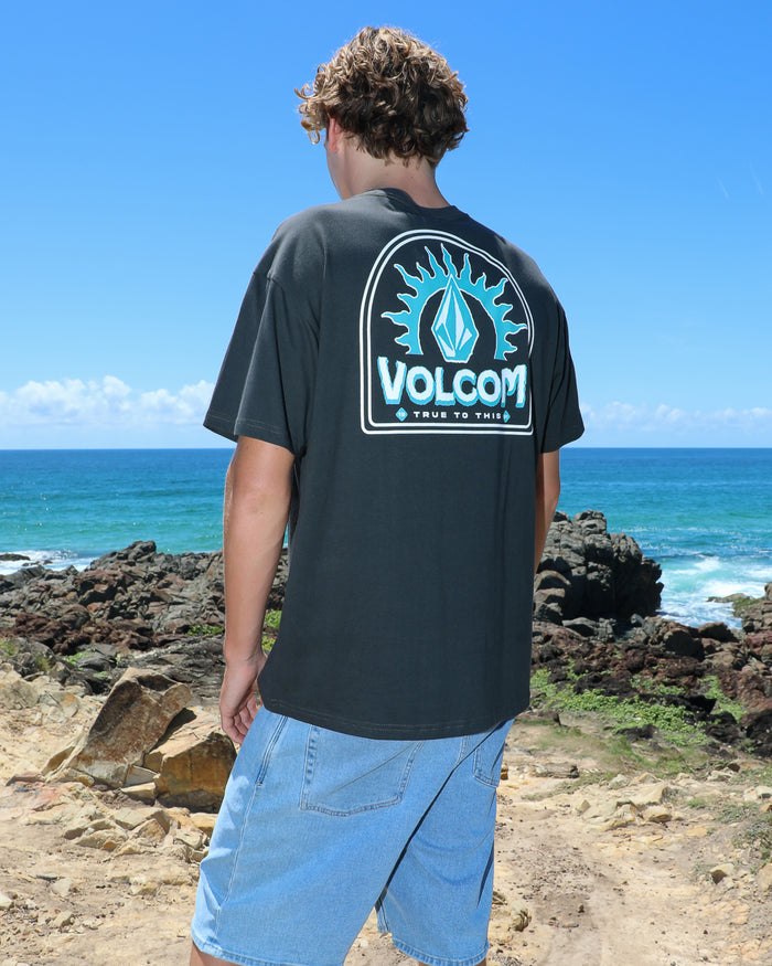 A person with curly hair and sunglasses, wearing a modern fit black Volcom Men's Solstice Short Sleeve Tee and denim shorts, stands on a rocky coastline overlooking the ocean beneath a clear blue sky.
