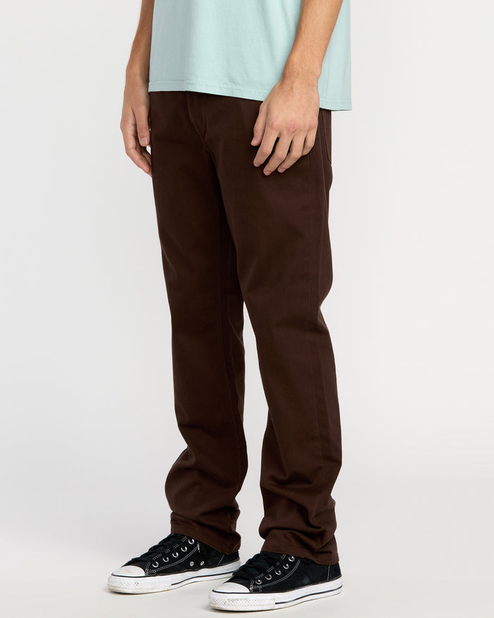 Wearing Volcom Men's Solver 5 Pocket Slub Pants, a light blue T-shirt, and black sneakers, a person stands against a plain white background. The cropped image highlights the stylish denim pants from the chest down.