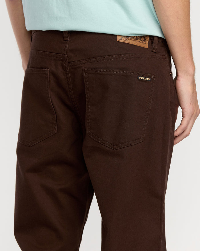 A man wears Volcom Men's Solver 5 Pocket Slub Pants in brown, featuring a visible logo on one back pocket. The image shows him from the waist down with one arm at his side.