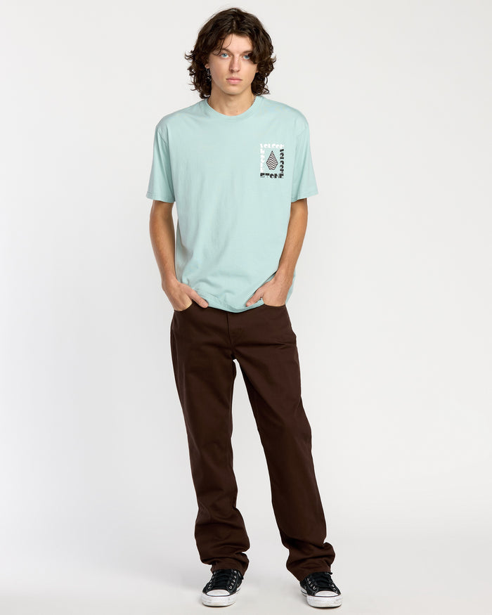 A person with medium-length curly hair faces forward, wearing a light blue Volcom T-shirt, Volcom Men's Solver 5 Pocket Slub Pants in brown, and black-and-white sneakers against a plain white background.