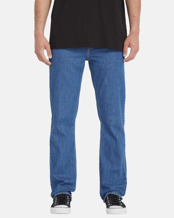 A person wearing a black t-shirt, Volcom Men's Solver Denim Pants in blue, and black-and-white sneakers, shown from the shoulders down against a plain white background.