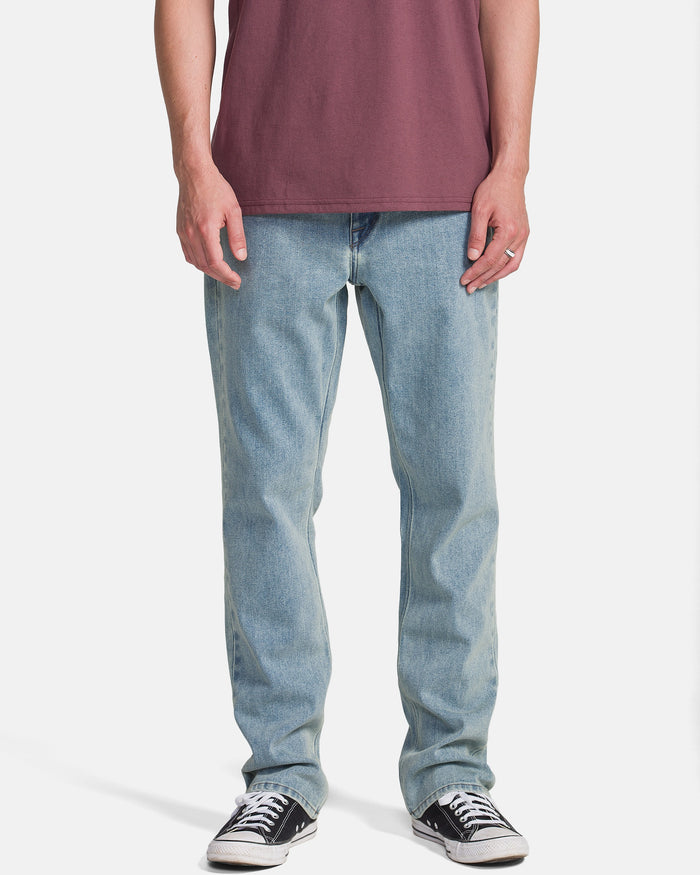 A person wearing a maroon t-shirt, Volcom Men's Solver Denim Pants in Dust Bowl Indigo, and black low-top sneakers stands against a plain white background. The image captures them from shoulders to feet.