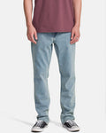 A person wearing a maroon t-shirt, Volcom Men's Solver Denim Pants in Dust Bowl Indigo, and black low-top sneakers stands against a plain white background. The image captures them from shoulders to feet.