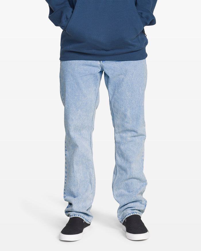 A person stands against a white background wearing a dark blue hoodie, black slip-on shoes, and Volcom Men's Solver Denim Pants in a bleached-out indigo wash. The upper body is partially out of the frame.