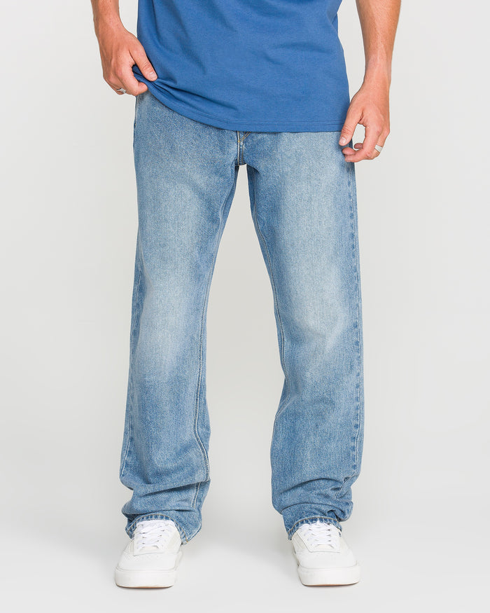 A person wearing a Freedom Blue T-shirt, Volcom Men's Solver Denim Pants in light blue stretch denim with a modern straight leg cut, and white sneakers stands facing forward against a plain white background. Their head is not visible in the image.