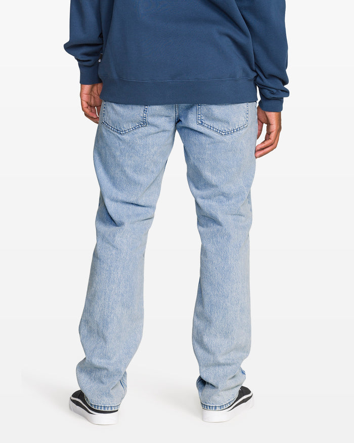 A person wearing Volcom Men's Solver Denim Pants in a bleached indigo wash, a navy sweatshirt, and black-and-white sneakers stands facing away from the camera against a plain white background.
