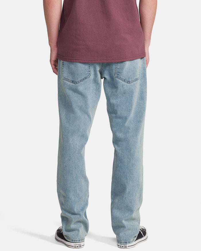 A person stands with their back to the camera, wearing Volcom Men's Solver Denim Pants in Dust Bowl Indigo, a maroon t-shirt, and black-and-white sneakers against a white background.