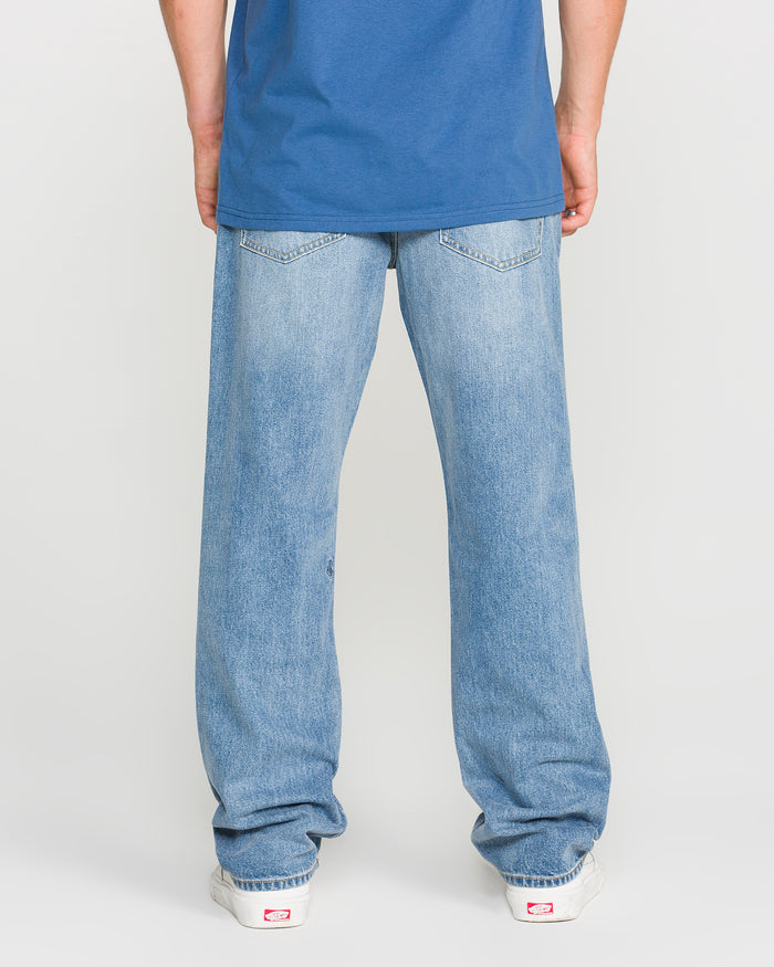A person stands facing away, wearing a blue t-shirt, white sneakers, and Volcom Men's Solver Denim Pants in Freedom Blue—modern straight-leg jeans in stretchy light blue denim. The background is plain and light-colored.