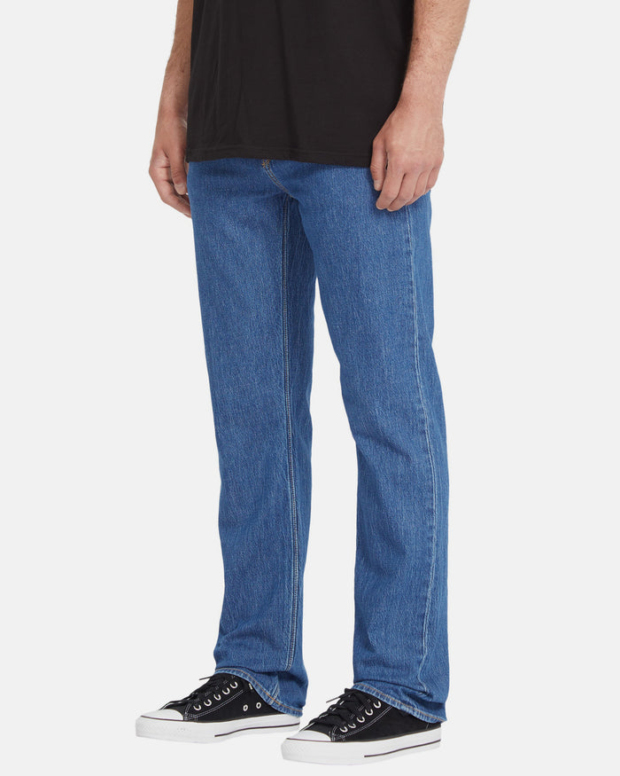 A person in Volcom Men's Solver Denim Pants—renowned for their Water Aware design—paired with a black t-shirt and black low-top sneakers with white soles, stands against a plain white background.