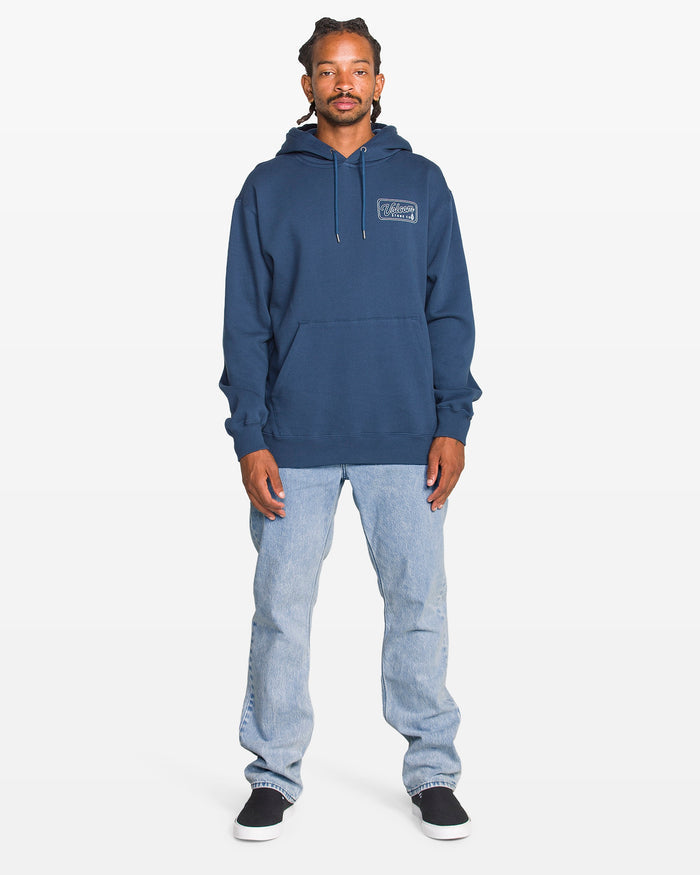 A man stands facing forward against a plain white background, wearing a blue hoodie, Volcom Men's Solver Denim Pants in a bleached-out indigo wash, and black slip-on shoes.