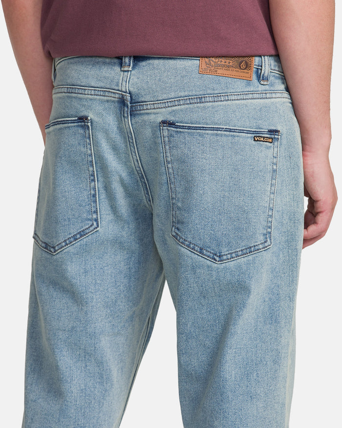 A person is shown from the back wearing Men's Solver Denim Pants by Volcom in Dust Bowl Indigo with a maroon shirt. The jeans feature two large back pockets and a brown Volcom patch on the waistband.